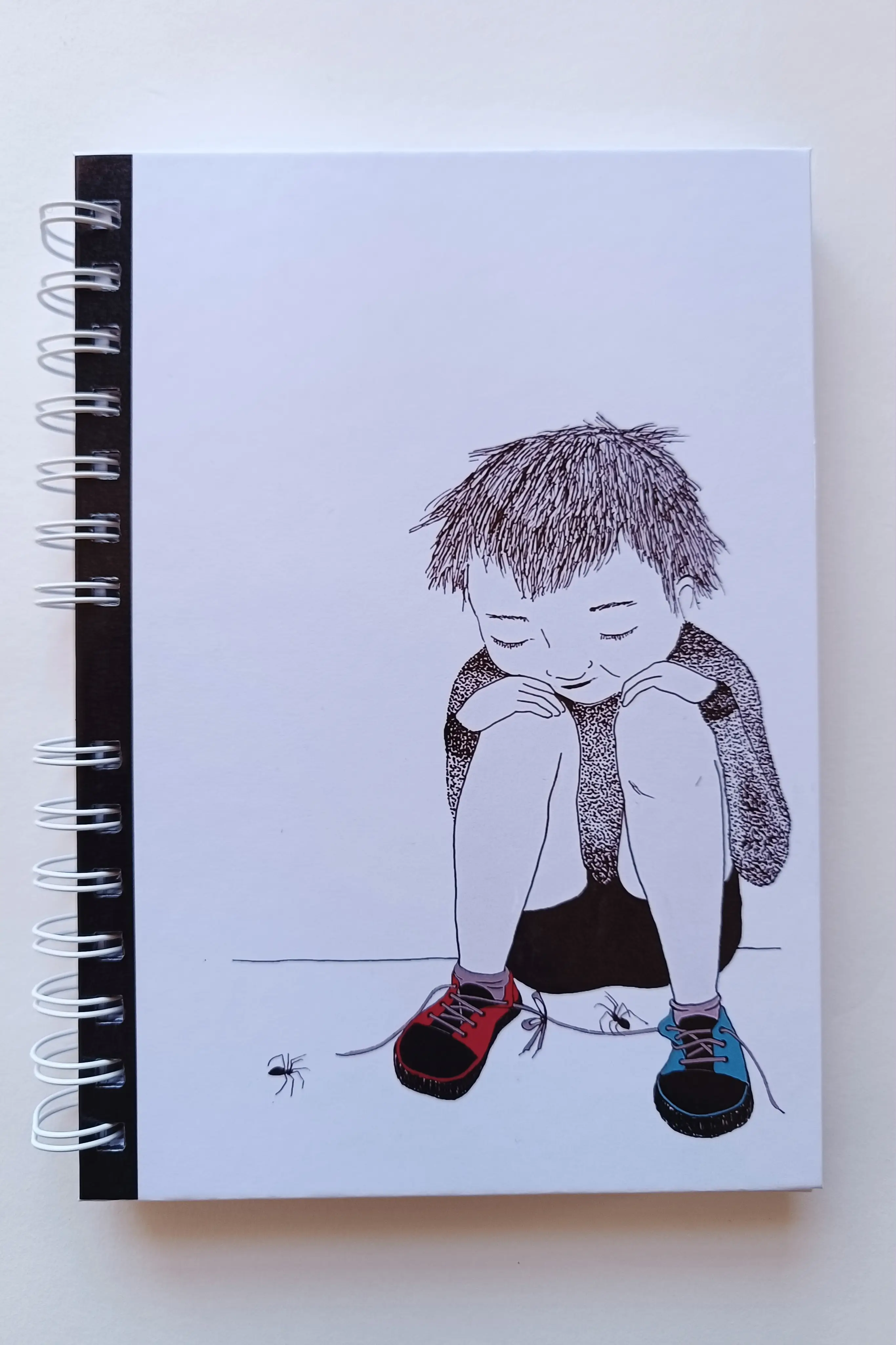Portada de libreta tamaño A5, con fondo blanco e ilustración a tinta que representa un niño sentado en el suelo con dos arañas